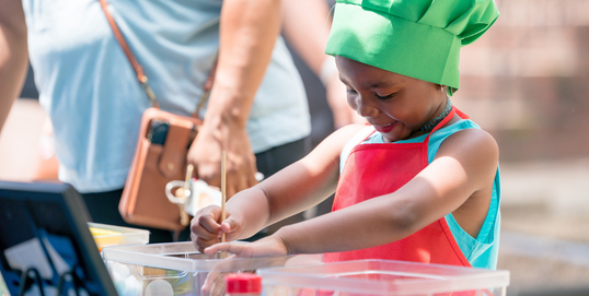 Young child with chef apron and hat with hands in a bowl of ingredients