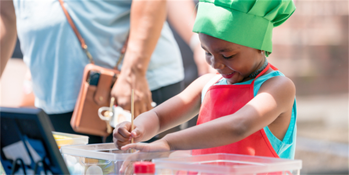 Young child with chef apron and hat with hands in a bowl of ingredients